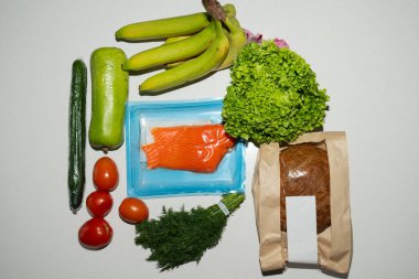 An assortment of fresh groceries, including ripe bananas, a head of lettuce, tomatoes, salmon and bread are laid out on a white surface.