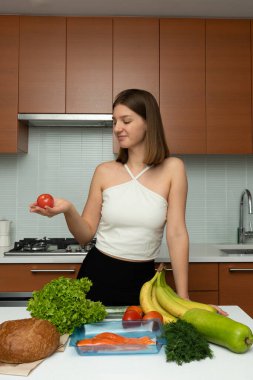 Modern mutfağında elinde tek bir domates tutan genç bir kadın var. Önünde beyaz tezgahta, sağlıklı bir yemek için yiyecek aranjmanı var..
