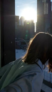 Balkondan güneşin doğuşunu izleyen bir kadın. Balkondan bakan kişi arkadaşlarını fark eder ve onlara gülümseyerek el sallar. Dışarıdaki işlek sanayi şehri görüntüsü - yavaş çekim, 4K Dikey Video