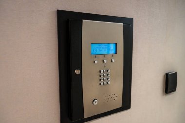 Duvara monte edilmiş, entegre dijital ekranı olan elektronik bir kasa. Kasa, siyah ve gümüş tonlarını birleştiren ve hem tuş takımı hem de anahtar erişimine sahip modern bir tasarıma sahip..