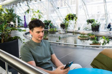 Bir adam cam bir avluda oturur, telefonuna bakar, etrafı saksı bitkileri ve göletle çevrili bir avluda..