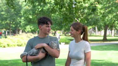 Genç bir adam, genç bir kadın ona yaklaşırken gri bir İngiliz Shorthair kedisini koşum takımı ile tutuyor. Halk parkında birlikte konuşup gülümsüyorlar. Gerçek zamanlı..