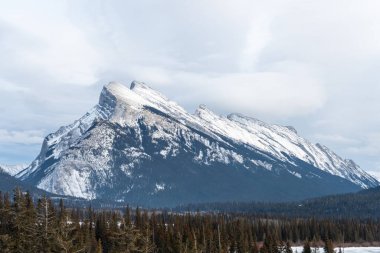 Manzarada, bulutlu bir gökyüzünün altında vadide ormanlar olan karlı bir dağ var. Seyahat, doğa, macera ve açık hava kampanyaları için idealdir.