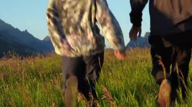 Güneşin doğuşuyla yemyeşil bir dağ çayırında el ele yürüyen iki insan. Waterton, AB, Kanada
