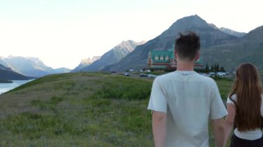 Waterton, AB, Kanada - 22 Ekim 2025: Bir çift, sakin gölü ve dağları olan büyük bir otele doğru çim tarlasından el ele yürür. Waterton, AB, Kanada