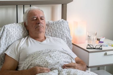 Yaşlanan yaşlı bir erkek, evde yatağında yorgun ve yorgun yatıyor. Tek başına dinlenen bir virüs. Mutsuz, üzgün bir şekilde uyuyor.