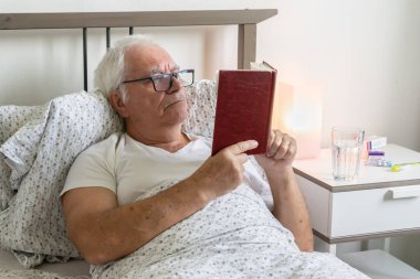 Yaşlanan yaşlı bir erkek, evde yatağında yorgun ve yorgun yatıyor. Tek başına dinleniyor. Virüsü tedavi ediyor. Mutsuz, üzgün, elinde kitap tutuyor.