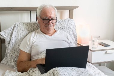 Yatakta üst düzey bir erkek. Ev ofisinde çalışıyor. Emekliler için dizüstü bilgisayar işleri yapıyor.