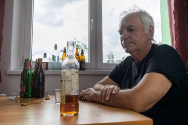 Yaşlı bir erkek masada oturur evde yalnız başına alkol şişesi içer üzgün alkolizm belirtileri ve rehabilitasyon sorunları ve iyileşme sorunları yaşar.