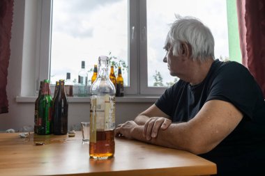 Yaşlı bir erkek masada oturur evde yalnız başına alkol şişesi içer üzgün alkolizm belirtileri ve rehabilitasyon sorunları ve iyileşme sorunları yaşar.