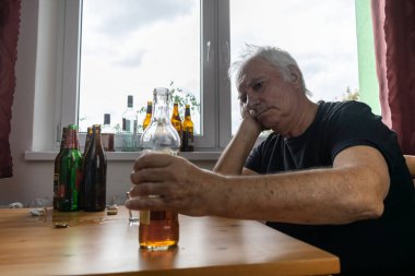Yaşlı bir erkek masada oturur evde yalnız başına alkol şişesi içer üzgün alkolizm belirtileri ve rehabilitasyon sorunları ve iyileşme sorunları yaşar.