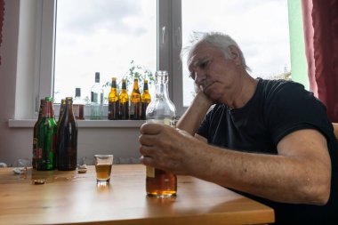 Yaşlı bir erkek masada oturur evde yalnız başına alkol şişesi içer üzgün alkolizm belirtileri ve rehabilitasyon sorunları ve iyileşme sorunları yaşar.