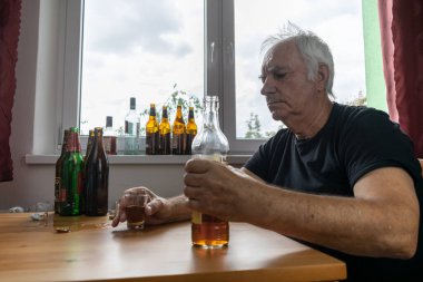 Yaşlı bir erkek masada oturur evde yalnız başına alkol şişesi içer üzgün alkolizm belirtileri ve rehabilitasyon sorunları ve iyileşme sorunları yaşar.