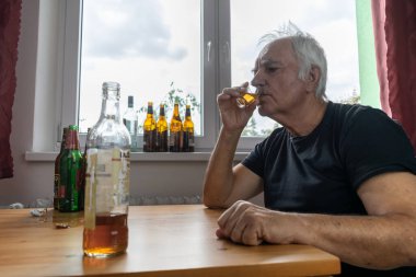 Yaşlı bir erkek masada oturur evde yalnız başına alkol şişesi içer üzgün alkolizm belirtileri ve rehabilitasyon sorunları ve iyileşme sorunları yaşar.