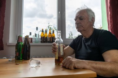 Yaşlı bir erkek masada oturur evde yalnız başına alkol şişesi içer üzgün alkolizm belirtileri ve rehabilitasyon sorunları ve iyileşme sorunları yaşar.