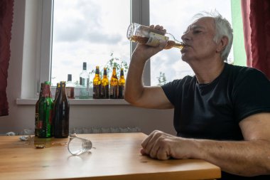 Yaşlı bir erkek masada oturur evde yalnız başına alkol şişesi içer üzgün alkolizm belirtileri ve rehabilitasyon sorunları ve iyileşme sorunları yaşar.