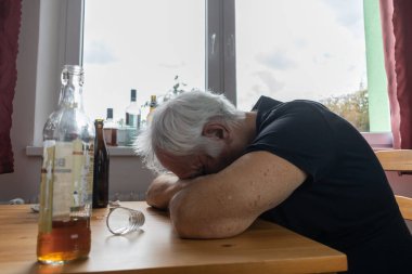 Yaşlı bir erkek masada oturur evde yalnız başına alkol şişesi içer üzgün alkolizm belirtileri ve rehabilitasyon sorunları ve iyileşme sorunları yaşar.