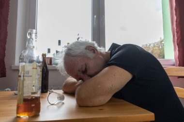 Yaşlı bir erkek masada oturur evde yalnız başına alkol şişesi içer üzgün alkolizm belirtileri ve rehabilitasyon sorunları ve iyileşme sorunları yaşar.