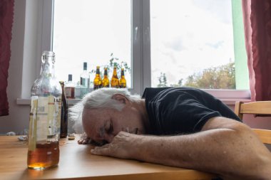 Yaşlı bir erkek masada oturur evde yalnız başına alkol şişesi içer üzgün alkolizm belirtileri ve rehabilitasyon sorunları ve iyileşme sorunları yaşar.