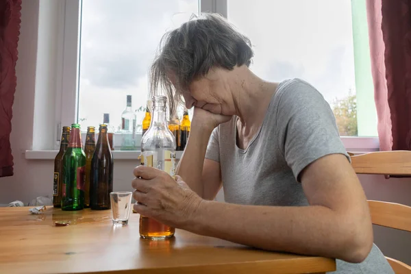 old senior female woman sit next to table drink alcohol bottle at home sad alone alcoholism Signs and Symptoms rehab abuse and recovery problems