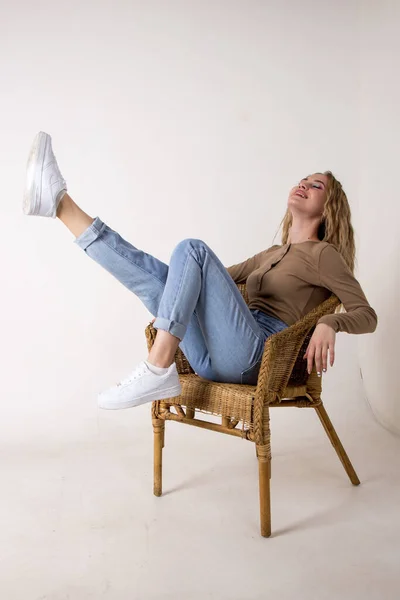 portrait of a beautiful young woman in jeans, shirt and sneakers.she put her feet up on the wicker rocking chair against the white brick wall.sitting on a chair.