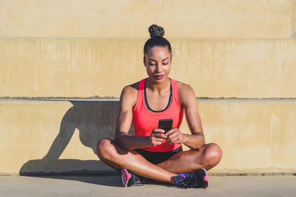 Çekici genç Afro-Amerikalı kadın saçları bağlanmış ve spor kıyafetleri giyiyor beton merdivenlerde bağlı oturuyor güneşli bir günde cep telefonunu kullanıyor ve gölge düşürüyor.