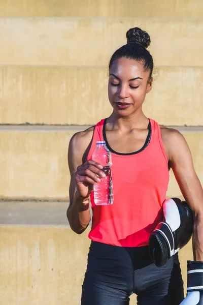 Güzel ve atletik bir Afro-Amerikan kadının portresi. Spor giysileri giymiş, boks eldiveni ve plastik bir şişe su tutuyor.