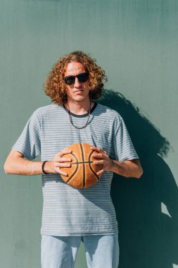 Elinde basketbol topu ve yeşil bir arka planı olan kameraya bakan adam.