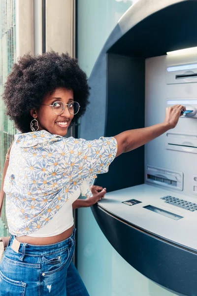 Kameraya bakıp gülümseyen Afrikalı genç bir kadın ATM kullanıyor.