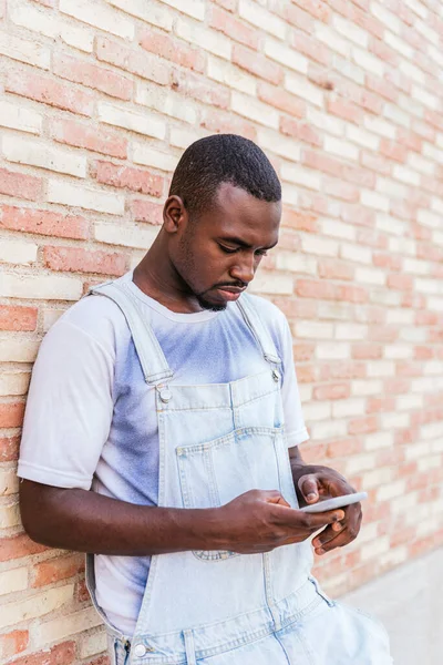 Молодий африканський чоловік спирається на цегляну стіну і використовує свій смартфон — стокове фото
