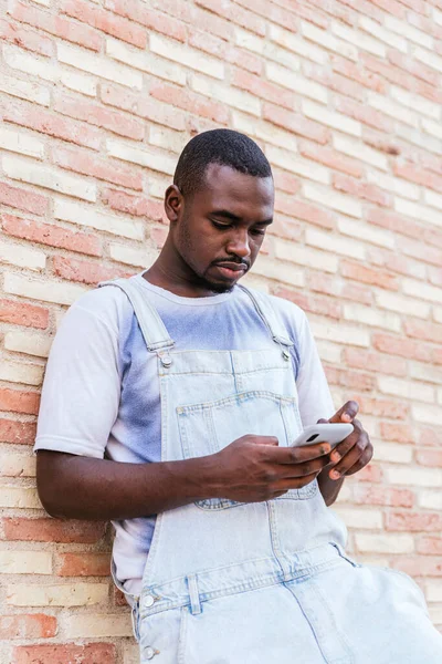 Молодий африканський чоловік спирається на цегляну стіну і використовує свій смартфон — стокове фото