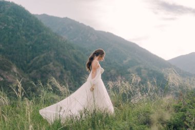 Gelinlik giymiş bir gelin, dağların arkasında bir tarlada yürüyor.