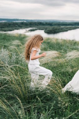 Beyaz elbiseli uzun saçlı bir kız nehir kıyısındaki tüy çimlerinde koşuyor.
