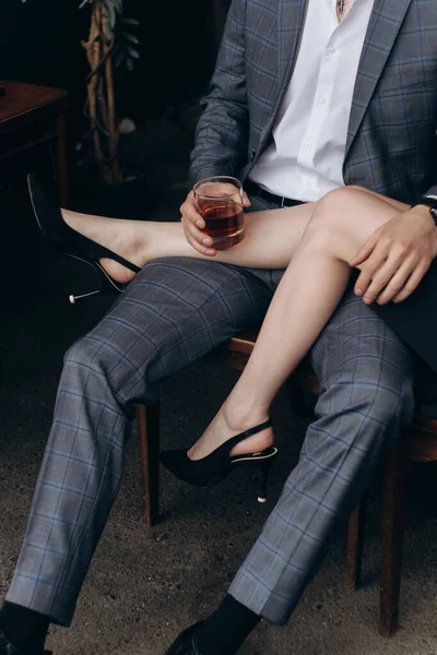 Kız, elinde bardak olan takım elbiseli bir adamın bacaklarının üzerine bacaklarını attı.
