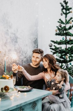 Çiçek, çelenk, maytap ve Noel ağacının yanında bir pastayla servis edilen bir masada kızı olan genç bir aile.