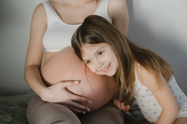 一个年轻的怀孕女孩 有一个女儿 穿着白色的家居服 住在一间明亮的房间里 — 图库照片