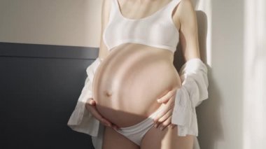 Beyaz iç çamaşırlı ve beyaz gömlekli hamile bir kız. Güneşte beyaz bir duvarın yanında.