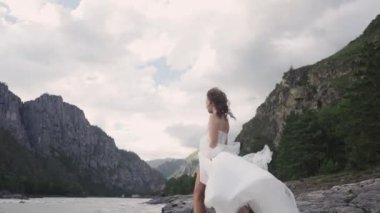 Gelinlikli bir gelin, Altai 'deki dağların arka planında, kayalık bir nehir kıyısında rüzgârda uçuyor.