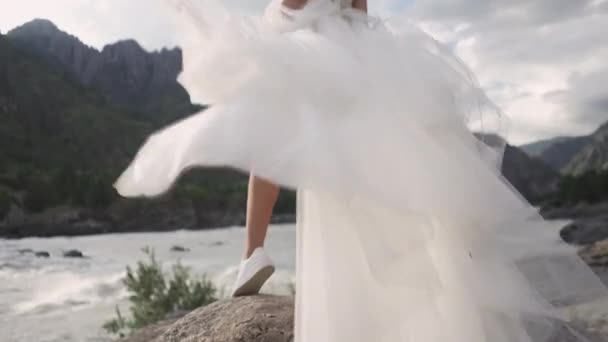 風になびくウェディングドレスを着た花嫁がアルタイの山を背景に岩の多い川岸に立っています — ストック動画