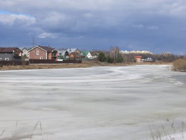 Köy göleti, uzak mesafede sıcak evleri olan bahar buzu kabuğuyla kaplı.