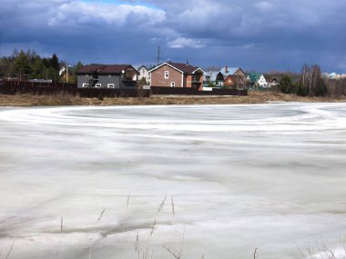 Köy göleti, uzak mesafede sıcak evleri olan bahar buzu kabuğuyla kaplı.