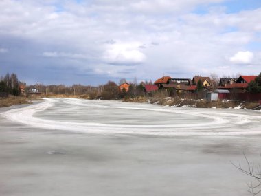 Köy göleti, uzak mesafede sıcak evleri olan bahar buzu kabuğuyla kaplı.