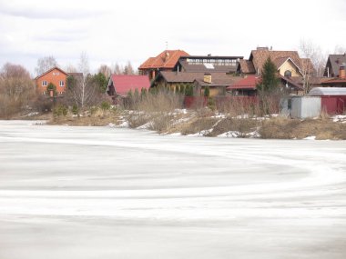 Köy göleti, uzak mesafede sıcak evleri olan bahar buzu kabuğuyla kaplı.