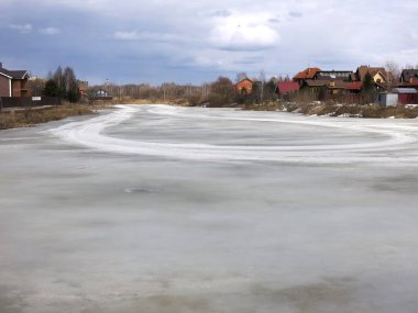 Köy göleti, uzak mesafede sıcak evleri olan bahar buzu kabuğuyla kaplı.