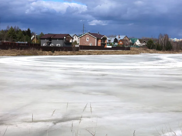 Köy göleti, uzak mesafede sıcak evleri olan bahar buzu kabuğuyla kaplı.