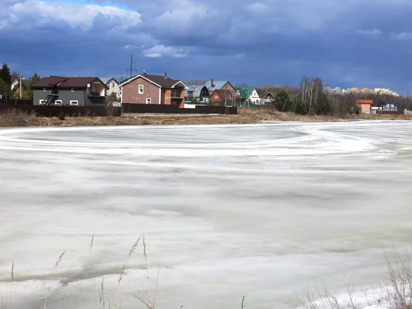 Köy göleti, uzak mesafede sıcak evleri olan bahar buzu kabuğuyla kaplı.