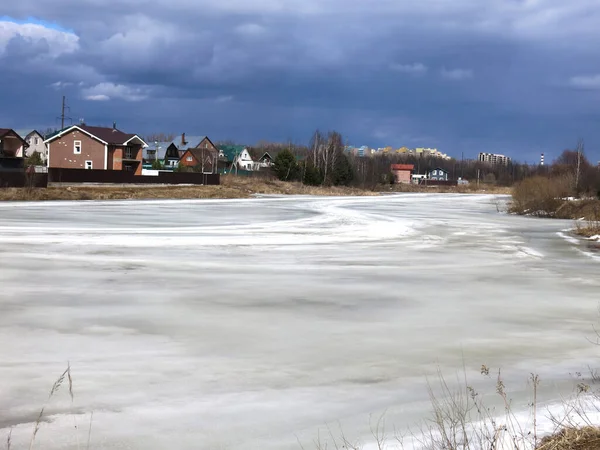 Köy göleti, uzak mesafede sıcak evleri olan bahar buzu kabuğuyla kaplı.