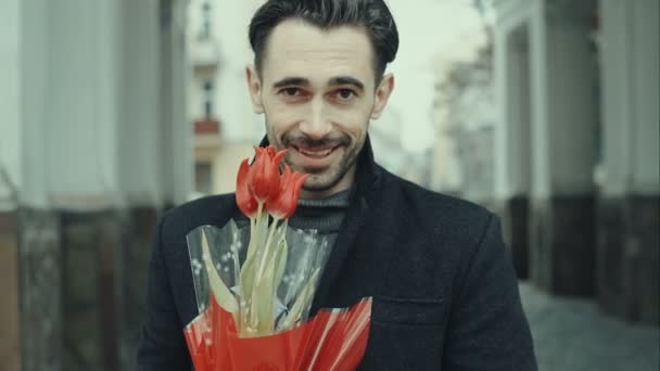 Happy smiling forty years old caucasian man with baguette and flower bouquet standing on the street. City buildings as background. RAW video rcord. — Stock Video