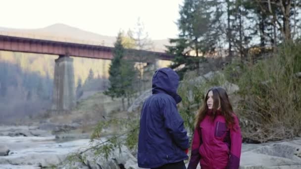 Turista chico y chica en la orilla de un río de montaña charla divertida — Vídeos de Stock