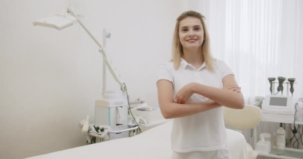 Hermosa joven cosmetóloga de pelo blanco Profesional en ropa blanca profesional de pie sonriente pone sus manos en el salón de belleza del hospital — Vídeo de stock